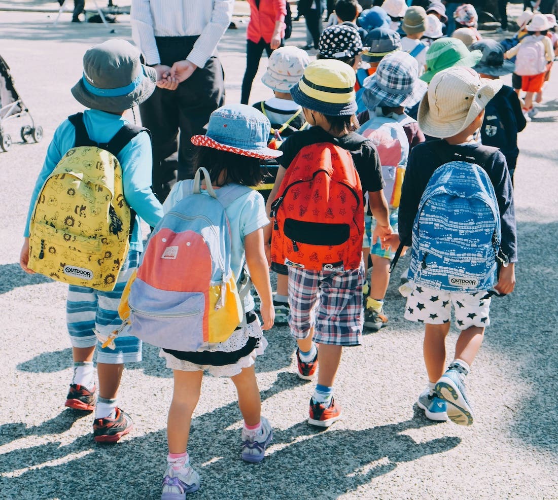 A medium shot photograph of a group of young children walking away from the viewer, their backs facing the camera. They are wearing colorful clothing and backpacks, and many have hats on. They appear to be walking in a line on a paved surface, possibly a sidewalk or courtyard, with a few adults visible in the background. The scene is brightly lit, suggesting daylight. The children’s clothing and the outdoor setting imply a casual, possibly school or outing-related context.